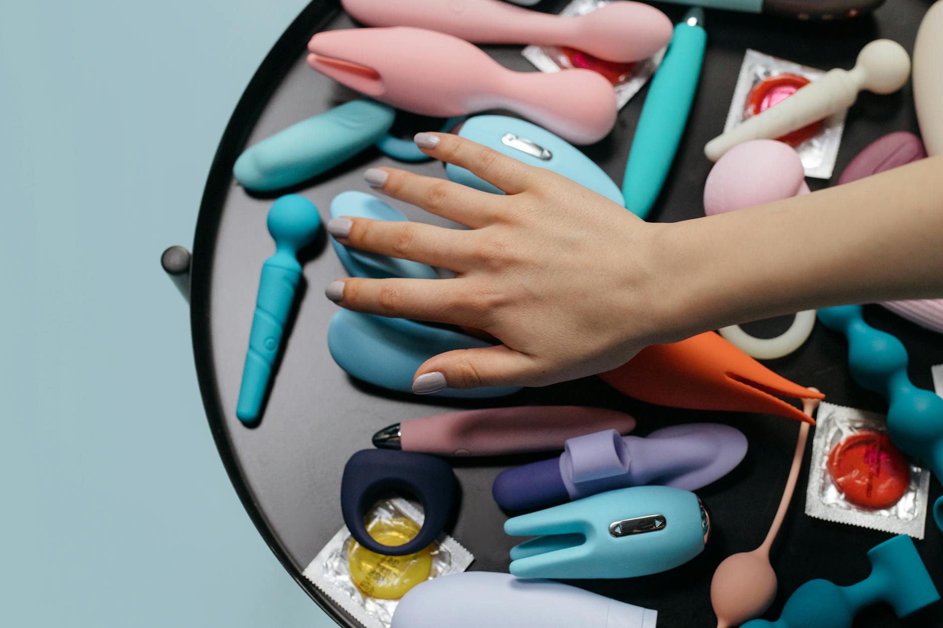 A hand reaching over a variety of colorful sex toys including a lemon vibrator arranged on a table.